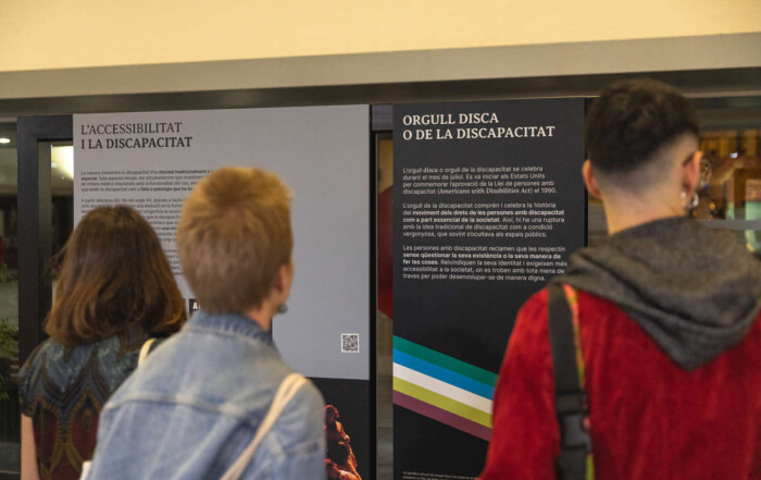 Tres persones dempeus, d'esquena a la càmera, i davant dos plafons de l'exposició "Obrir portes, aixecar telons. El què, qui, com de l'accessibilitat en arts escèniques" al vestíbul de l'Institut del Teatre.